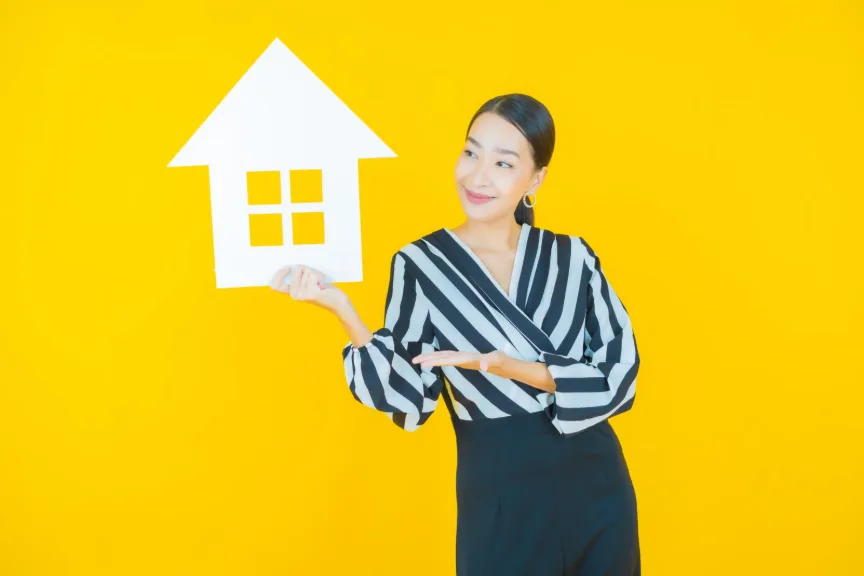 Smiling Asian woman holds paper house prop against yellow background, promoting Roomz rentals.