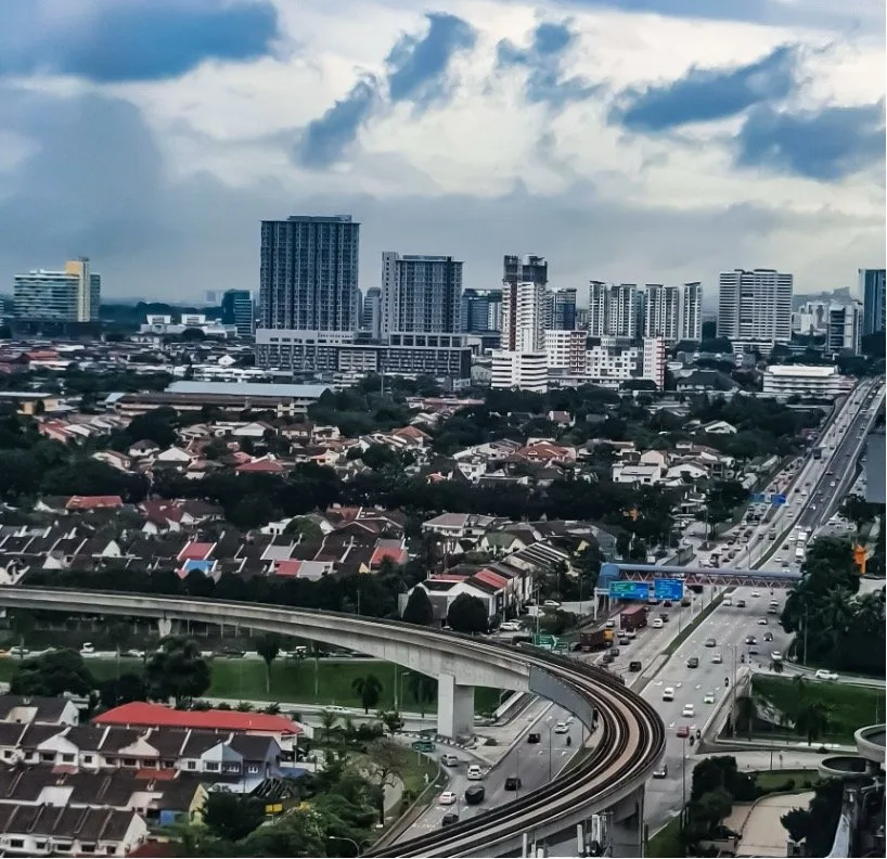 Aerial view of a modern city with high-rise apartment and office buildings in the background, a residential neighborhood with red-roof houses in the foreground, and an elevated rail line curving alongside a busy multi-lane highway under cloudy skies. Room